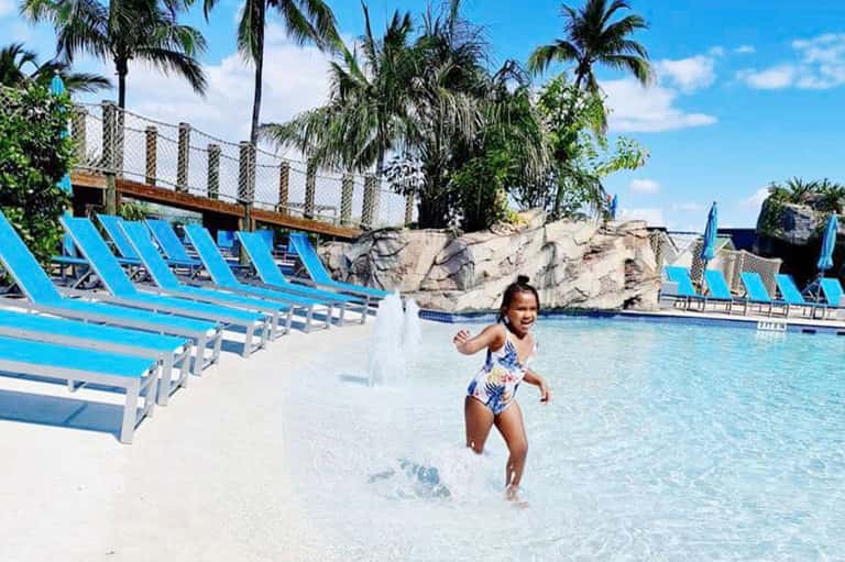 Child running into shallow pool water with blue lounge chairs and palm trees in background