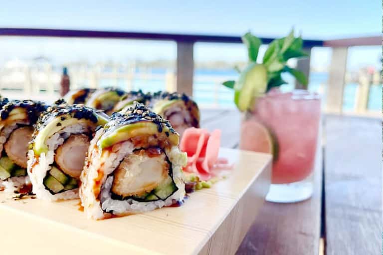 Sushi rolls on a wooden board with pink cocktails in the background overlooking the ocean