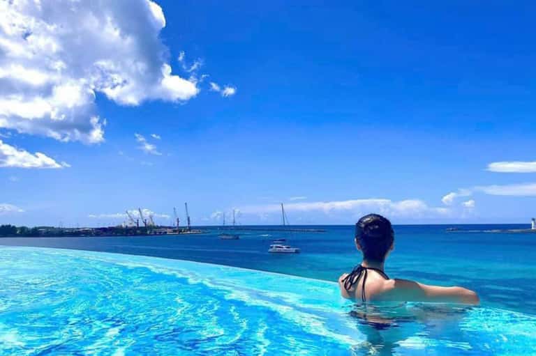Woman swimming in infinity pool overlooking turquoise ocean waters with boats and industrial port