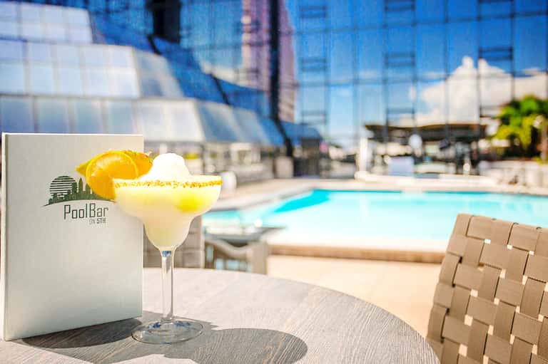 Tropical cocktail with orange slice on poolside table overlooking bright blue swimming pool and city buildings