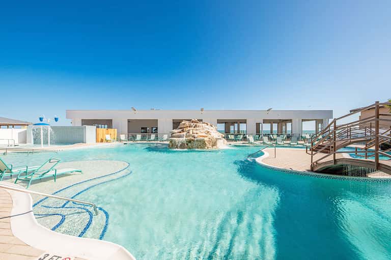 Turquoise resort pool with rock feature, loungers, and modern white building at daytime