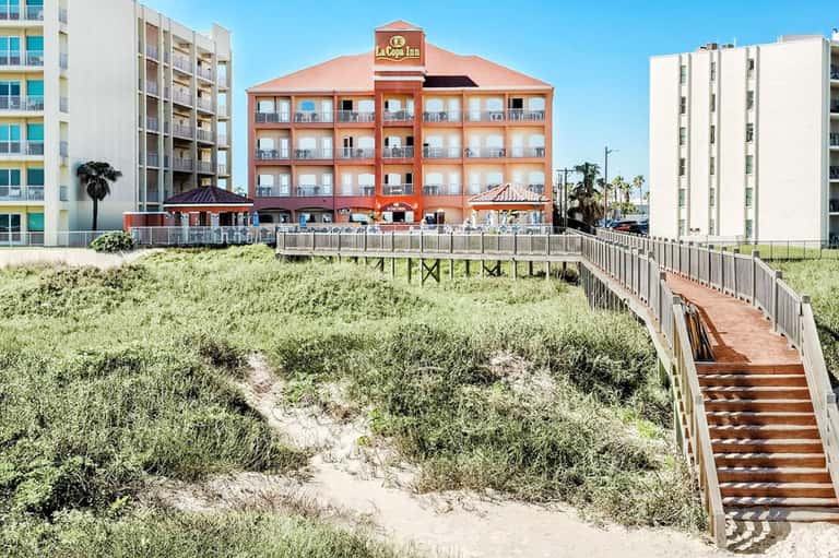 Beachfront hotel with red exterior, wooden boardwalk over dunes, and adjacent modern buildings