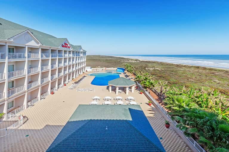 Beachfront resort with multi-story white building, blue pool, and ocean view beyond sandy dunes