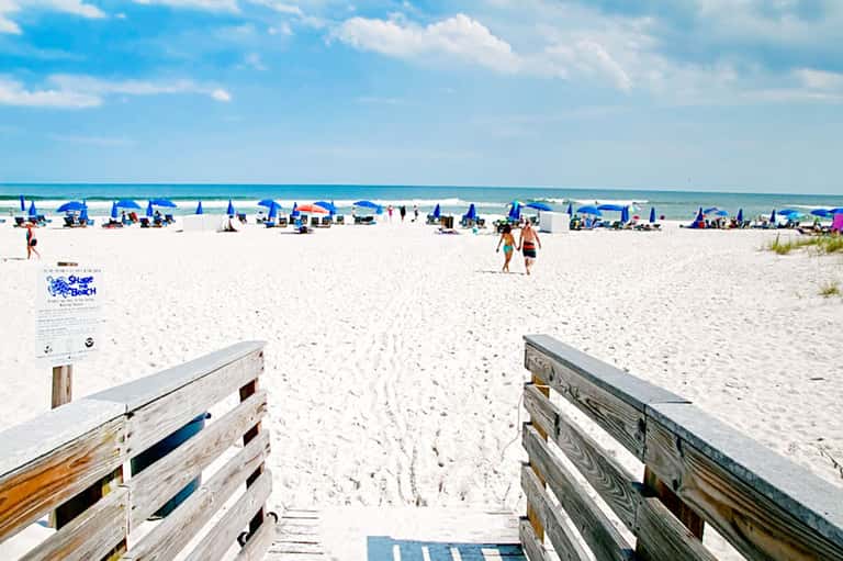 Sandy beach access with wooden boardwalk, blue umbrellas, and ocean under clear sky