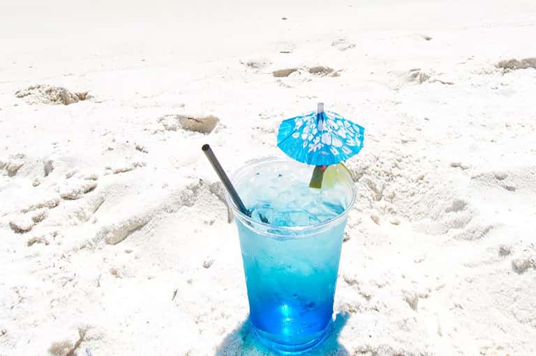 Tropical blue cocktail with umbrella and straw on sandy beach with footprints and ocean backdrop