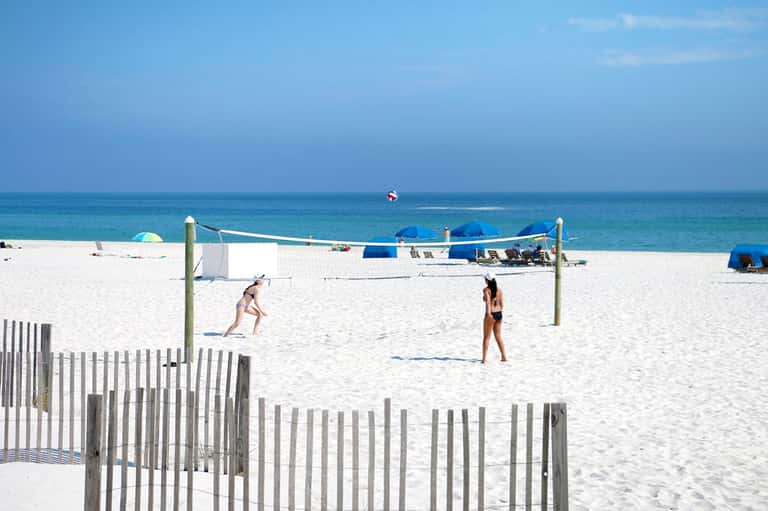 Beach volleyball court with players, ocean view, white sand, and blue umbrellas