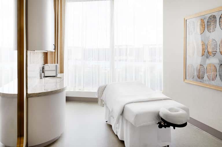 Minimalist spa treatment room with white massage bed, round sink, and floor-to-ceiling windows