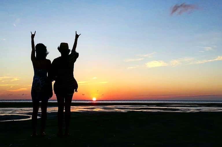 Two people silhouetted against sunset on beach with arms raised in celebration