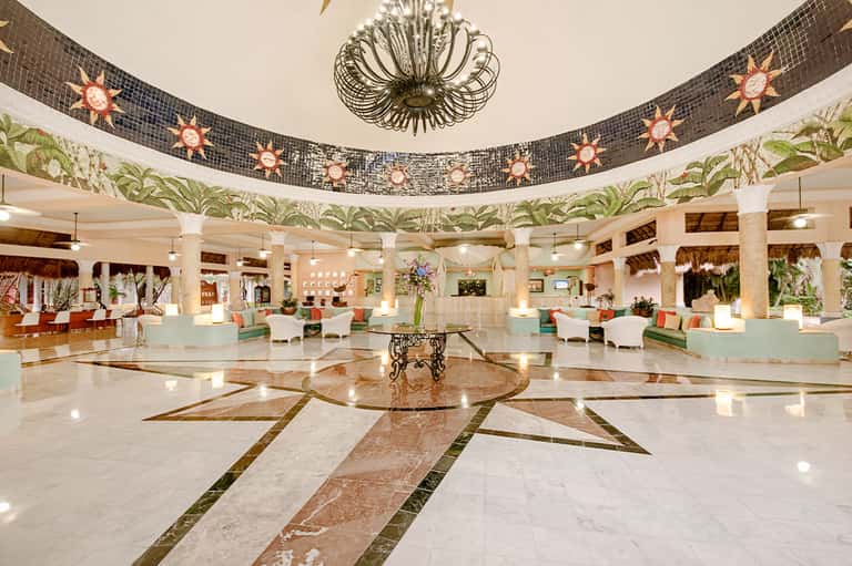 Grand hotel lobby with circular marble floor pattern, decorative sun ceiling medallions, and palm plant displays