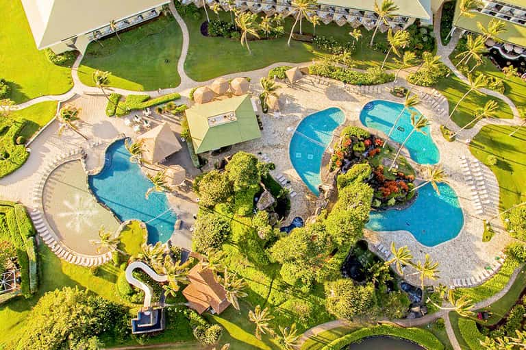 Aerial view of resort pool complex with curved blue pools surrounded by palm trees and manicured landscaping