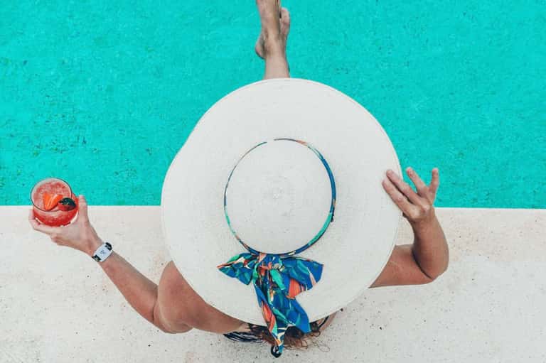 Person in turquoise water holding white sun hat and colorful drink, poolside shot