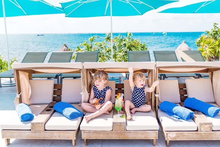 Two young children sitting in wooden cabana beds with ocean view and blue umbrellas at beachfront resort