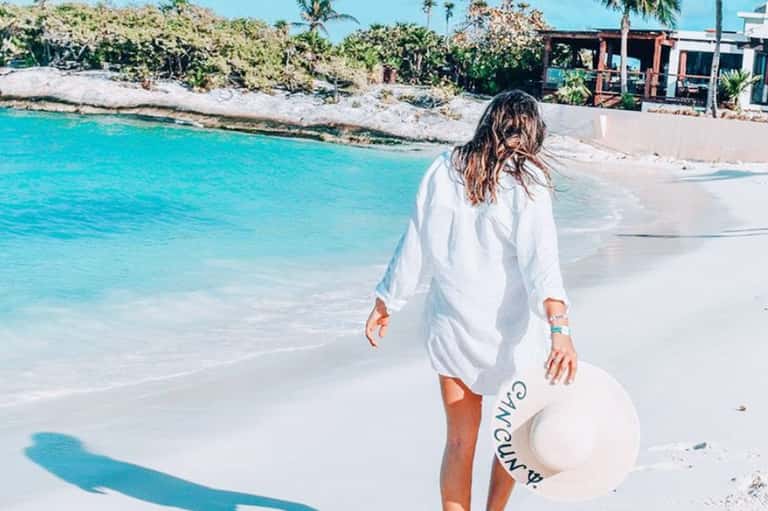 Woman in white shirt walking on white sand beach toward turquoise ocean water
