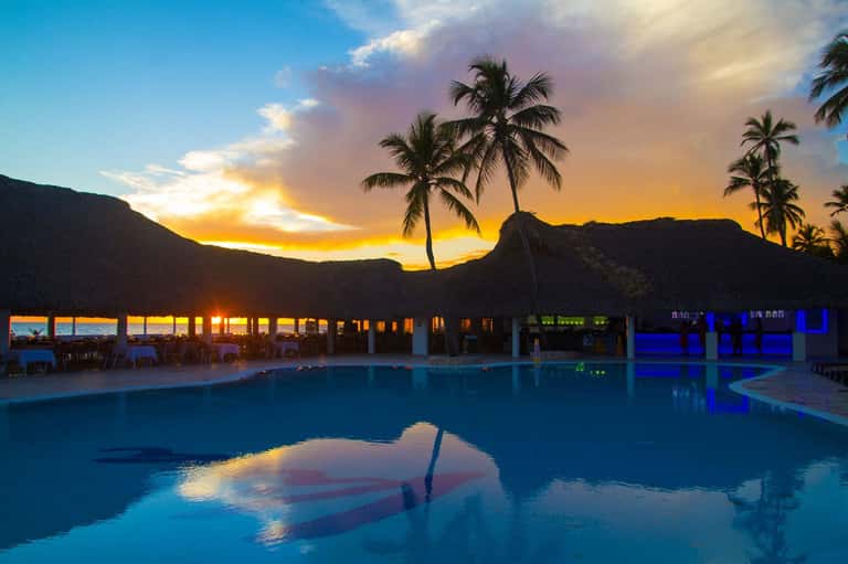 Resort pool at sunset with palm trees, illuminated dining pavilion, and mountains silhouetted against golden sky