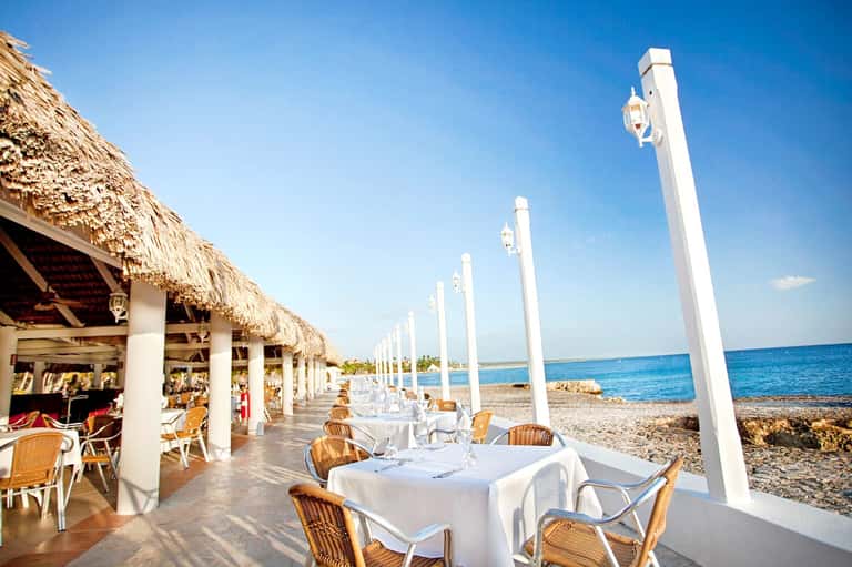 Beachfront restaurant terrace with white columns, rattan chairs, and thatched roof covering