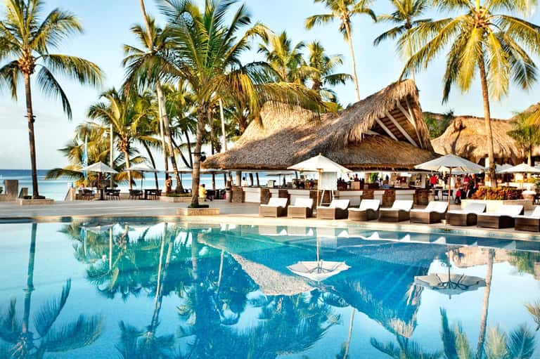 Tropical beachfront resort pool with thatched roof structures, palm trees, and ocean view