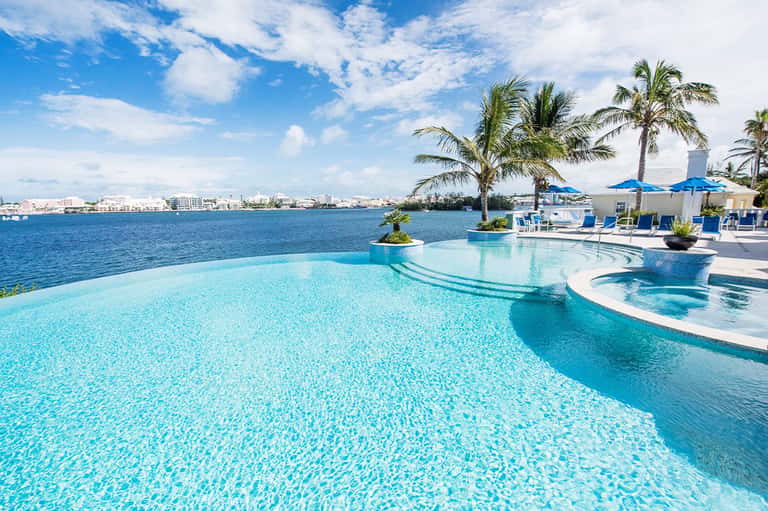 Luxury beachfront infinity pool overlooking ocean with palm trees, umbrellas, and coastal city skyline