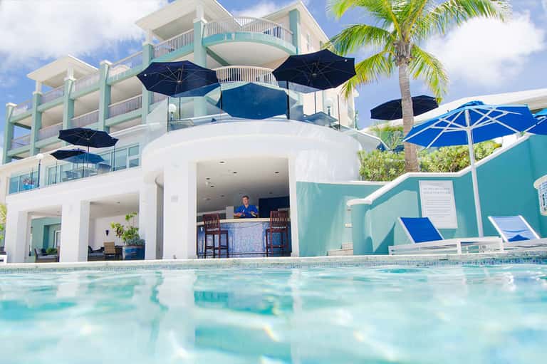 Resort pool with turquoise water, white building with blue umbrellas, palm trees, and staff at bar