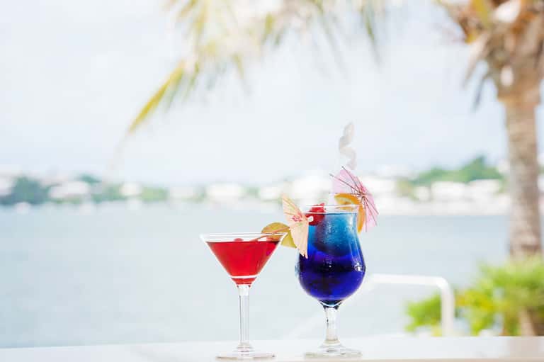 Two tropical cocktails with colorful umbrellas on table overlooking calm bay waters and palm trees