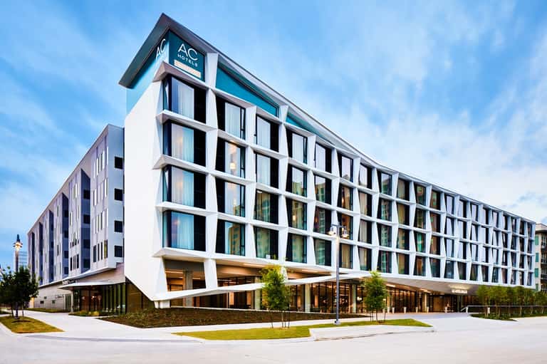 Modern AC Hotel exterior with white facade, grid-pattern windows, and ground-level entrance with landscaping