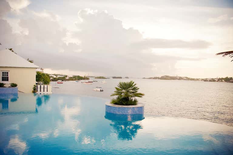 Infinity pool overlooking calm bay with sailboats, palm tree planter in pool, white building and cloudy sky