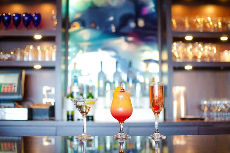 Three colorful cocktails displayed on bar counter with bottles in background