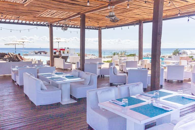 Beachfront lounge area with white furniture, wooden pergola, ocean view, and umbrella