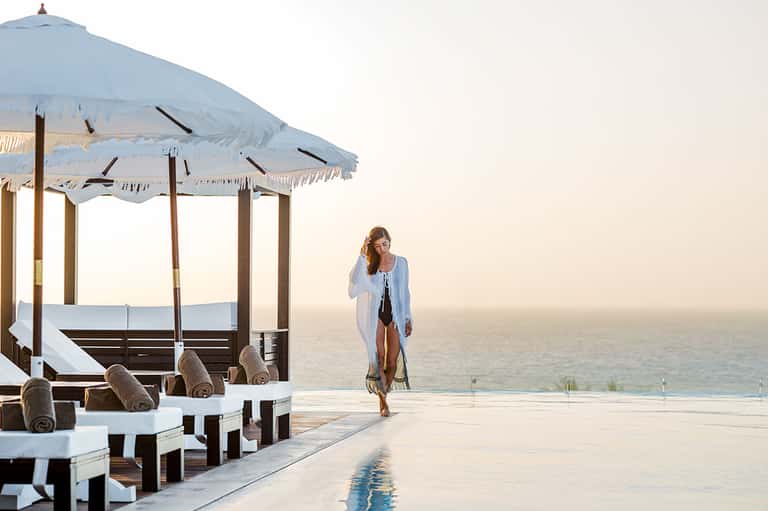 Woman in white cardigan walking by infinity pool overlooking calm ocean at dusk with blue umbrella cabana