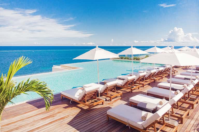 Infinity pool overlooking turquoise ocean with white umbrellas, wooden deck loungers, and tropical palm fronds