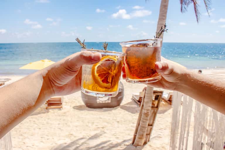 Two tropical rum cocktails with orange slices and palm fronds clinking glasses at beach with ocean view