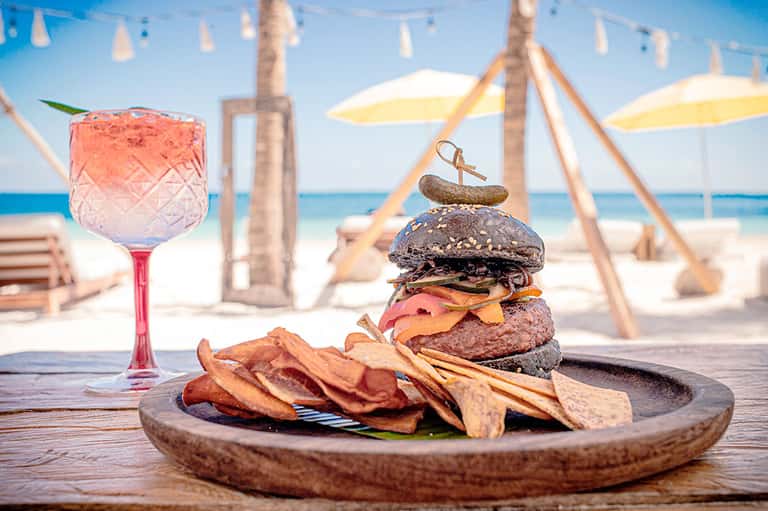 Gourmet burger and fries on wooden plate with colorful cocktail on beach with umbrellas and ocean backdrop