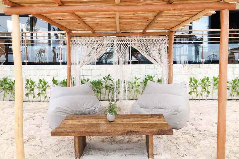 Beach cabana with wooden pergola, macramé curtains, white cushioned daybed, and ocean view
