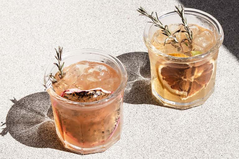Two iced cocktails with rosemary sprigs in glasses on sunny concrete patio surface