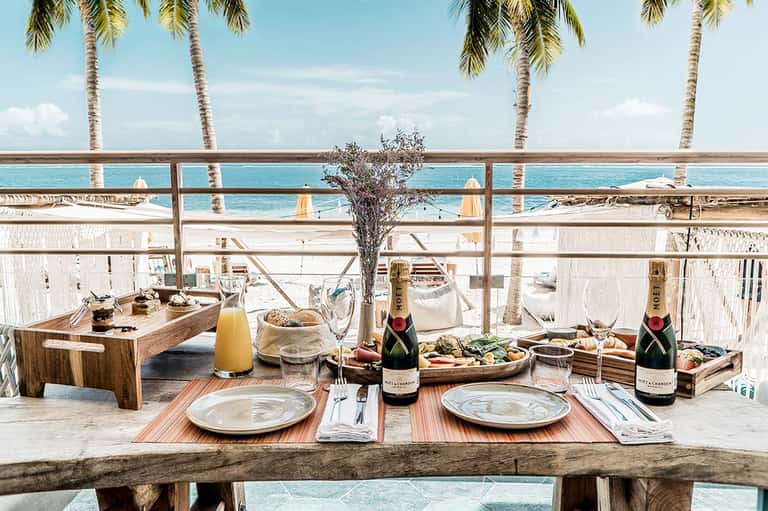 Beachfront dining table set for two with champagne bottles and ocean view framed by palm trees