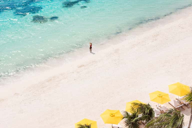 Aerial view of pristine beach with turquoise water, white sand, and yellow umbrellas with lone person wading