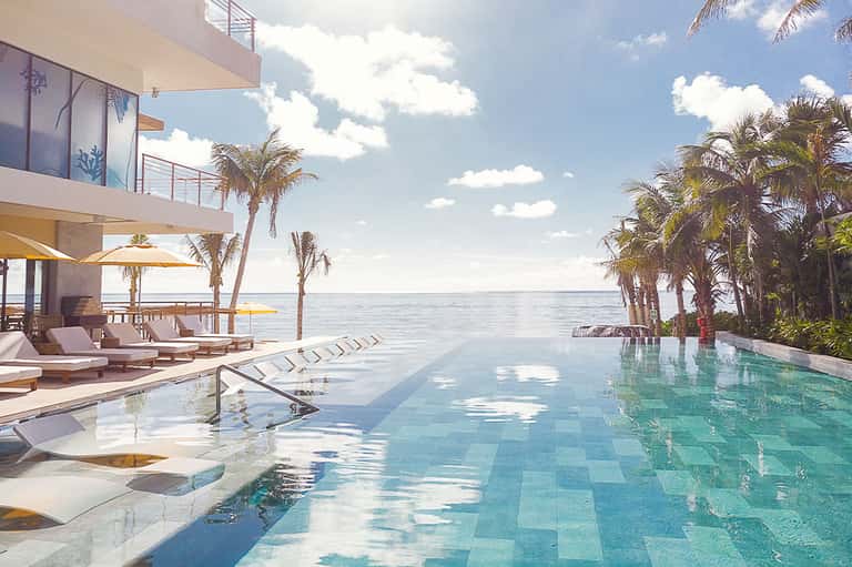Luxury infinity pool with ocean view, palm trees, lounge chairs, and modern beachfront architecture