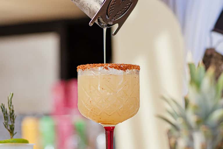 Cocktail being poured into a glass with spiced rim and red stem