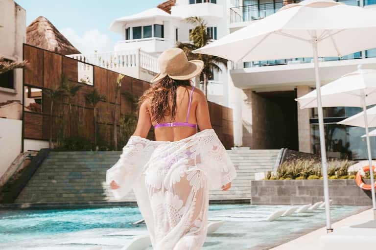Woman in swimsuit and straw hat walking toward pool area at beachfront resort with white umbrellas