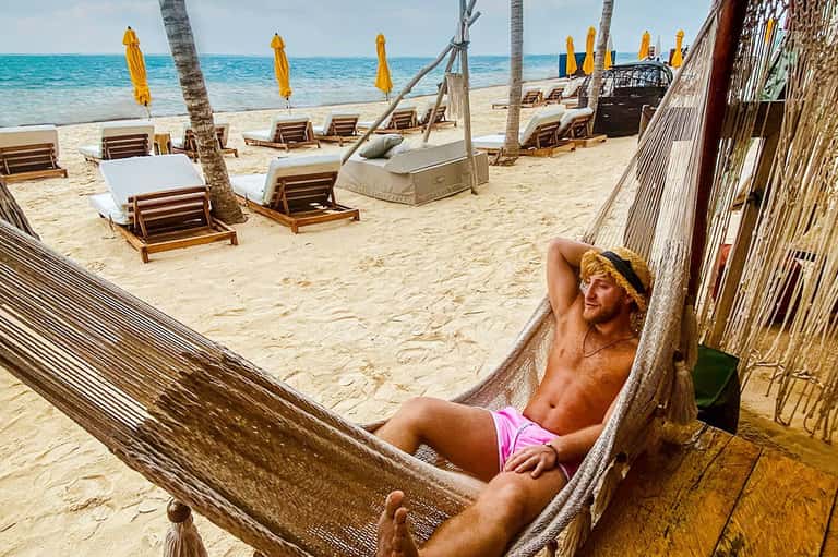 Man relaxing in hammock on tropical beach with palm trees, lounge chairs, yellow umbrellas, and turquoise ocean