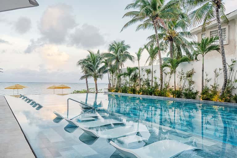 Infinity pool overlooking ocean with white loungers, palm trees, and white building architecture
