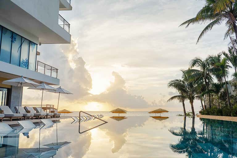 Luxury infinity pool at sunset with palm trees, modern building, golden sky reflections in still water