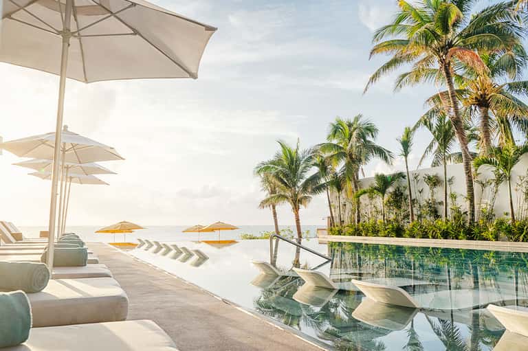 Oceanfront resort pool deck with white umbrellas, palm trees, and infinity pool overlooking the sea