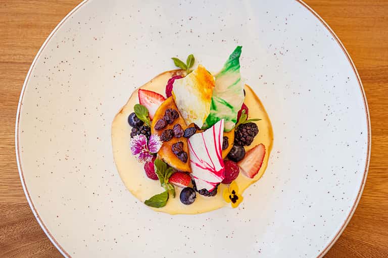 Artistic plated dessert with berries, edible flowers, and garnishes on white speckled plate