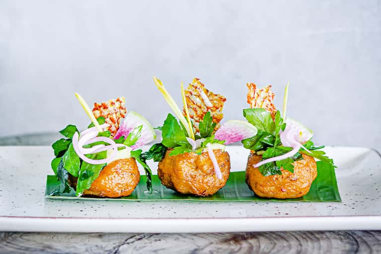 Gourmet appetizer plate with three golden fried items, pink onion garnish, and colorful flower decoration