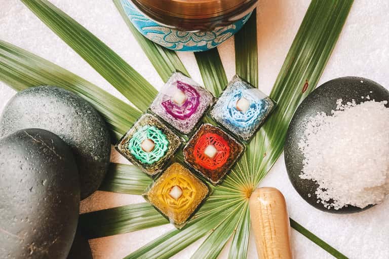 Spa massage stones, mortar and pestle, colorful paste bowls, and palm leaves arranged on white surface