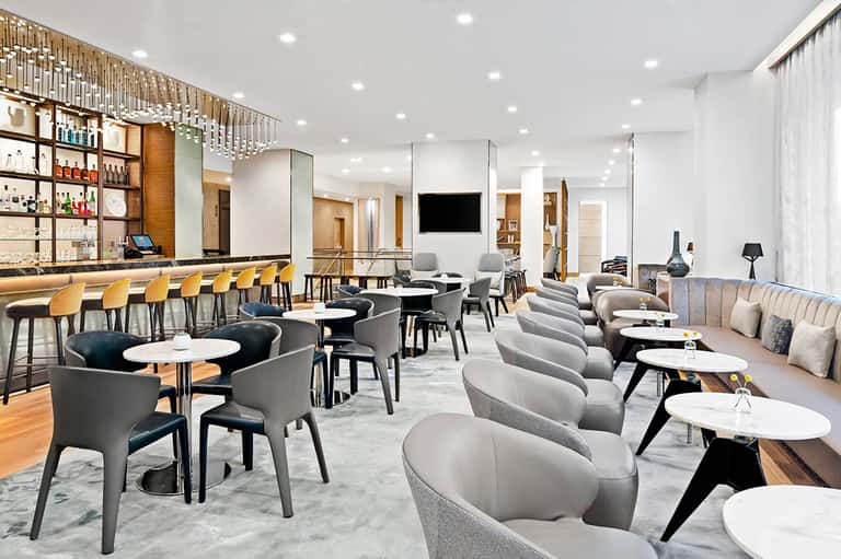 Modern hotel bar and lounge with gray seating, white tables, and wooden accents