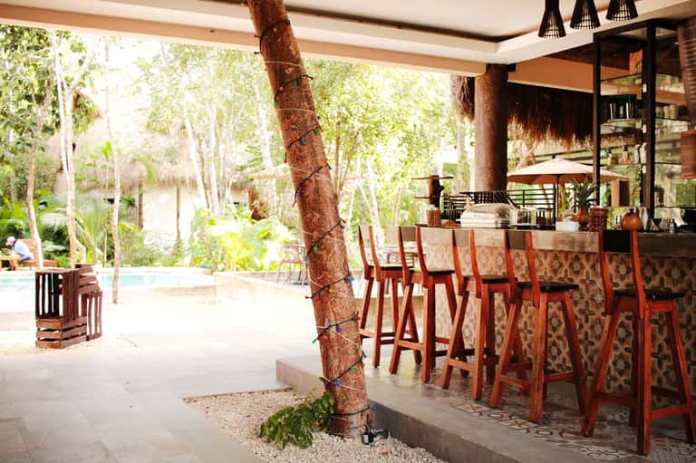 Open-air resort bar with wooden counter, tall chairs, and views of tropical grounds with trees