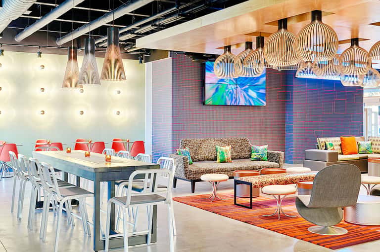 Modern hotel lounge with colorful pendant lights, dining table, patterned sofa, and digital display wall