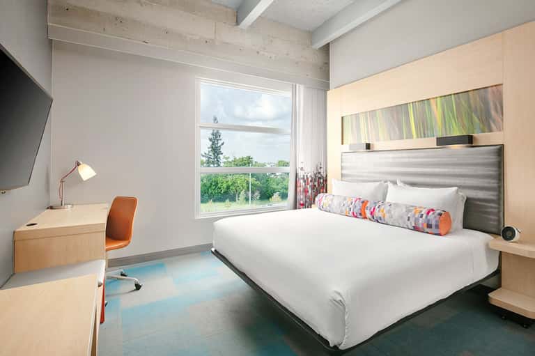 Modern hotel bedroom with queen bed, work desk, floor-to-ceiling windows, and contemporary furnishings