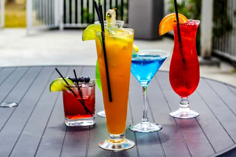 Four colorful cocktails on outdoor deck with lime and orange garnishes